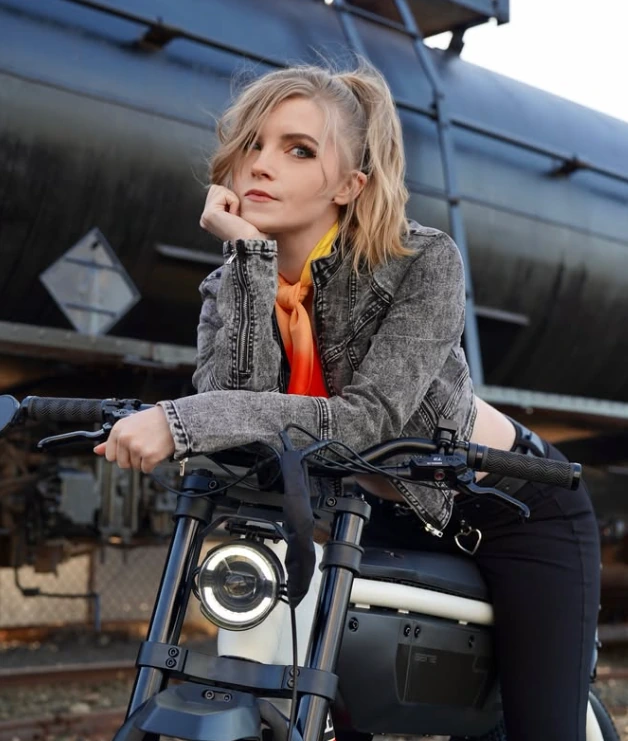Jessica Marie leaning on a motorcycle in a denim jacket and orange scarf